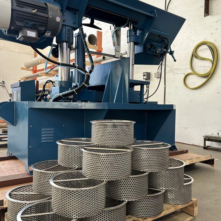 Pallet of perforated stainless steel strainer baskets beside a bandsaw in the DELC fabrication shop