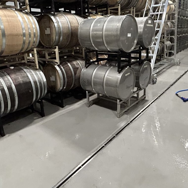 Stainless slot drain running down a winery barrel room floor lined with wine barrels